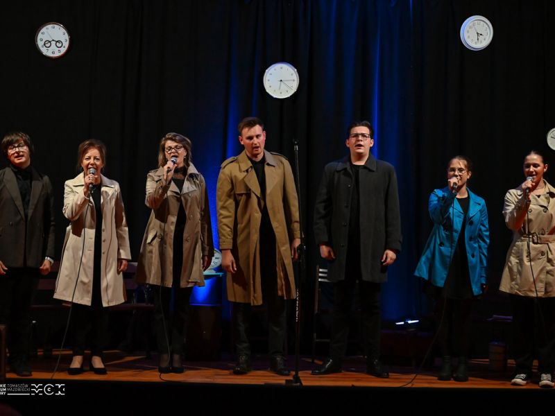 Na zdjęciu artyści Teatru na scenie, ubrani w jesienne płaszcze. Część z nich trzyma w w dłoniach mikrofony. Za nimi granatowa kurtyna, na której wiszą zegary.